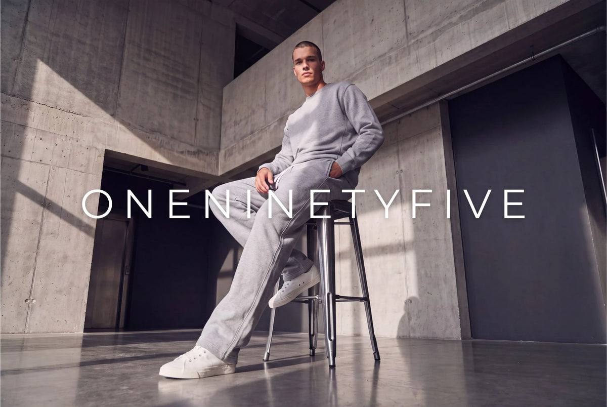 Model in grauem Sweatshirt und grauer Jogginghose von oneninetyfive sitzt auf einem Barhocker in einem Betonraum, davor der Schriftzug ONENINETYFIVE